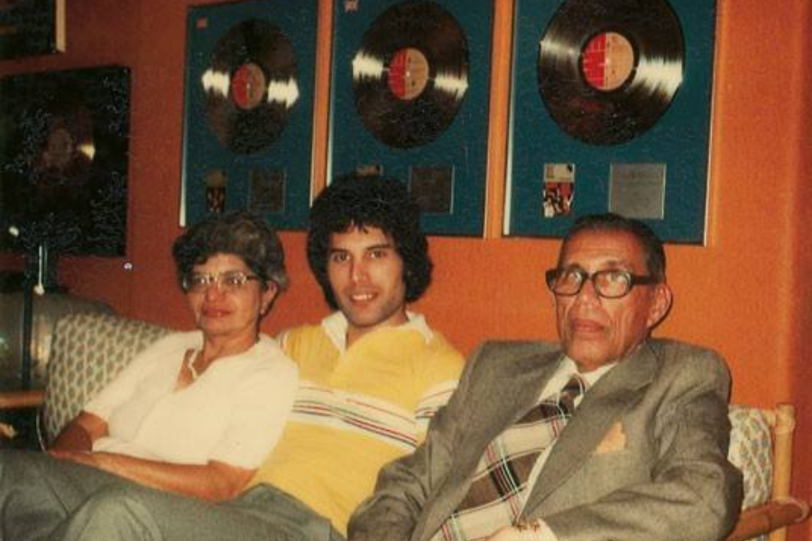 Color photograph of a casually dressed young man seated between an older woman and an older man in a cozy room with an orange wall. Framed gold or platinum records are mounted behind them, indicating musical success. The trio is relaxed and smiling gently, suggesting a family moment in a music-themed setting.