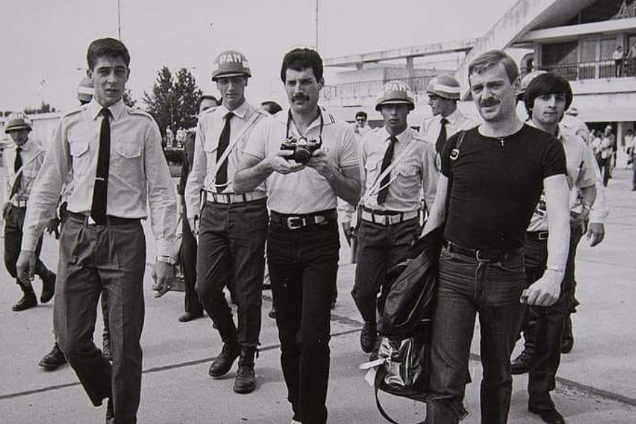 Black-and-white photo showing Freddie Mercury walking with a group of people, including uniformed security personnel and his partner Jim Hutton. Freddie holds a camera and wears a tucked-in polo shirt and black pants, while Jim is dressed in a fitted black tee and jeans, carrying a bag. The atmosphere appears to be that of an arrival or departure at a public venue, possibly an airport or stadium.