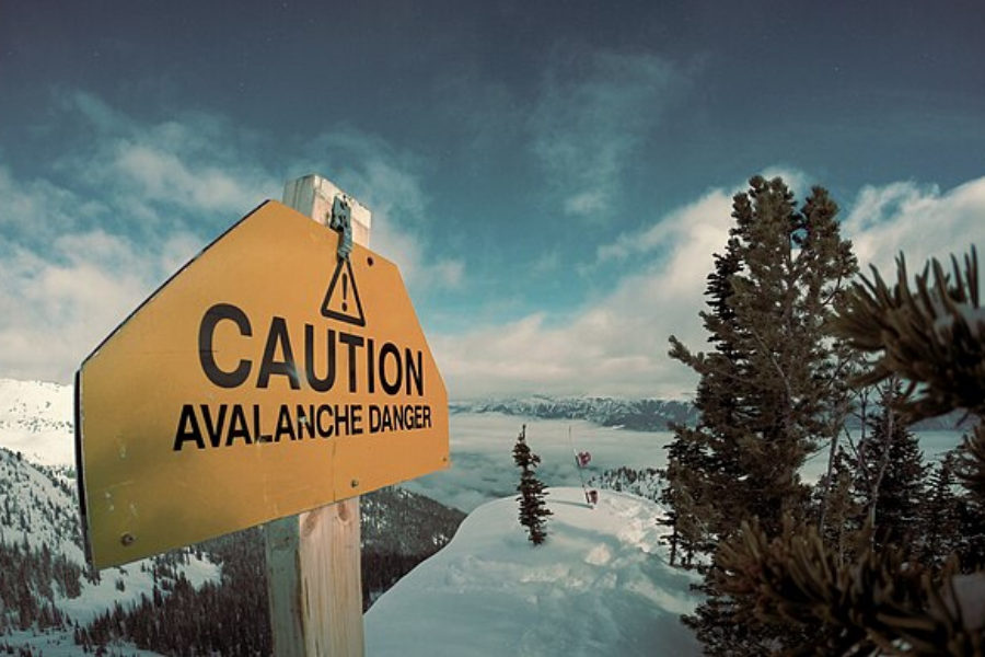 A bright yellow sign reading “CAUTION AVALANCHE DANGER” is posted on a snowy mountain ridge surrounded by evergreen trees. The dramatic view shows distant snow-covered peaks and a sky filled with clouds and sunlight.