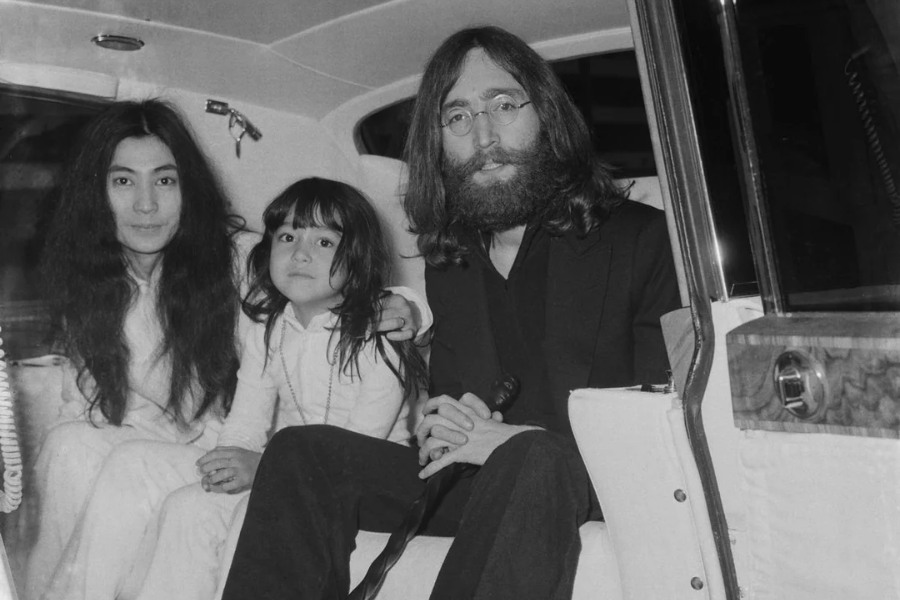Black-and-white photo of a woman, a young girl, and a bearded man with long hair and glasses, sitting together in the back of a car. The woman and girl are dressed in light clothing, while the man wears a dark suit and holds a cane, all looking toward the camera.