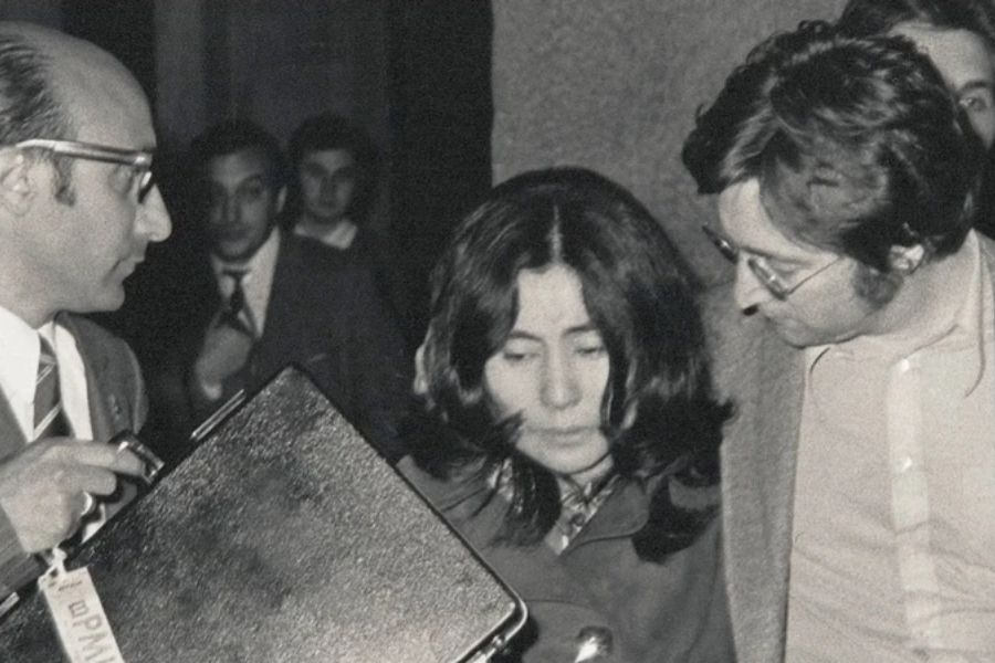 Black-and-white photo of a woman with long dark hair looking downward, surrounded closely by two men in suits and glasses. One man holds a briefcase, and several people are visible in the background, giving the scene a serious, tense atmosphere.