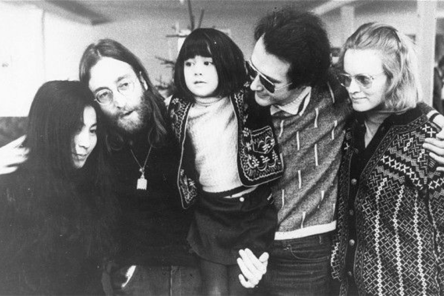 Black-and-white photo of two men, two women, and a child standing closely together indoors. The group is smiling or looking warmly at each other, with the child wearing a patterned jacket and sitting on one man's hip, while the adults wear winter sweaters.