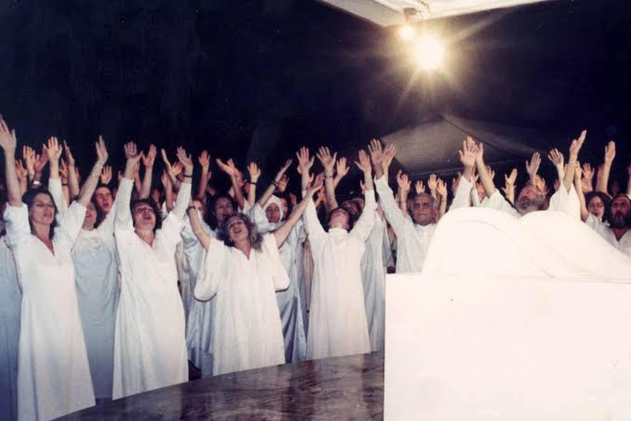 Color photograph of a large group of people dressed in white robes, standing under a bright light with their arms raised enthusiastically towards the sky. They appear to be participating in a group ritual or spiritual gathering, with dark surroundings and a white structure in the foreground.