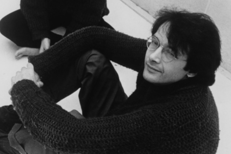 Black-and-white photo of a man with dark hair and glasses sitting on the floor with his arms wrapped around his knees. He wears a thick knitted sweater and looks up at the camera with a slight smile.