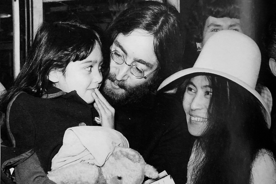 Black-and-white photo of a bearded man with glasses holding a young girl who touches his face affectionately, while a smiling woman in a wide-brimmed hat looks on. The child holds a stuffed animal, and the scene appears joyful and warm.