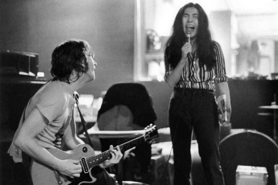 Black-and-white photo of a man playing an electric guitar while seated and looking up at a woman standing beside him, passionately singing into a microphone. The woman wears a striped shirt and jeans, and they appear to be rehearsing or performing in a casual indoor setting.