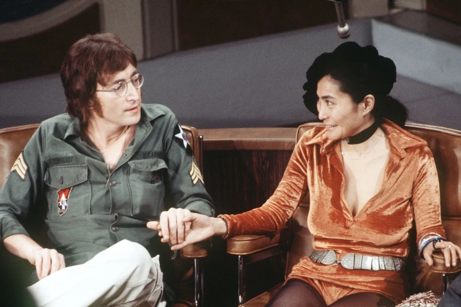 Color photograph of a man in a military-style green shirt and round glasses holding hands with a woman in a deep orange velvet mini dress and wide silver belt. They are seated on stage chairs during what appears to be a television interview, gazing at each other with smiles.