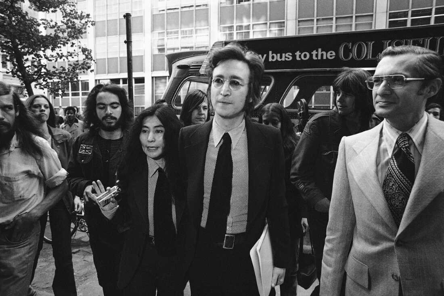 Black-and-white photo of a man and woman in matching suits and ties walking together in a crowd, accompanied by another man in a light-colored suit. They appear to be leaving or arriving at an event, with a bus and office buildings in the background.