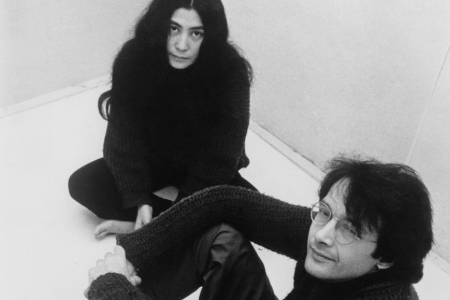 Black-and-white photograph of a woman with long dark hair sitting cross-legged on the floor beside a man with glasses and wavy hair, who leans back with his arms resting on his knees. Both wear dark, chunky sweaters and look towards the camera in a minimalist, light-colored room.