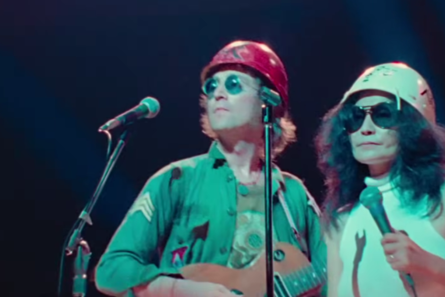 Color photograph of a man and woman on stage wearing hard hats and sunglasses. The man, dressed in a green military-style shirt, holds a small guitar while the woman, in a white sleeveless top, holds a microphone as they look out toward the audience.
