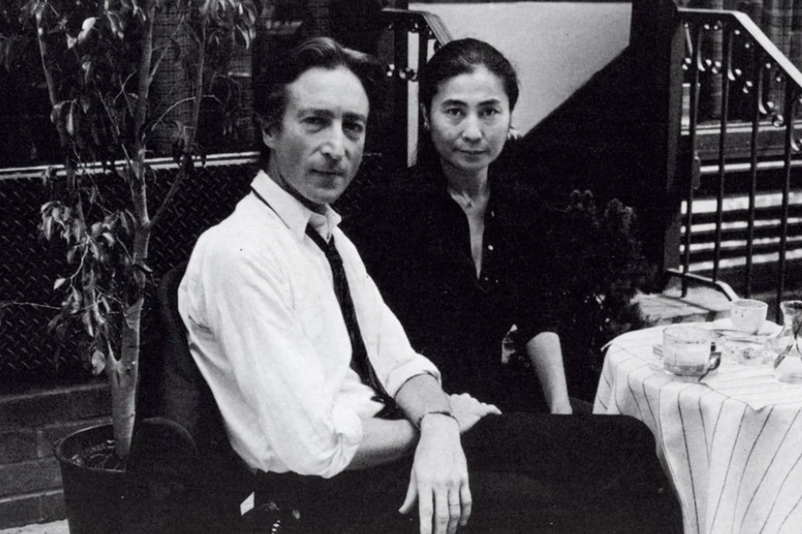 Black-and-white photo of a man in a white shirt and loosened tie seated at an outdoor café table beside a woman in a dark blouse. They both look directly at the camera with calm, composed expressions, surrounded by plants and a striped tablecloth with cups and glasses.
