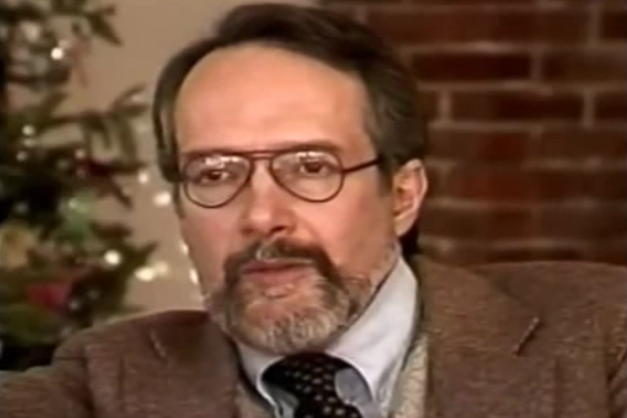 Color image of a man with glasses, a beard, and short hair, wearing a brown blazer, light shirt, sweater vest, and polka-dot tie. He appears to be speaking or mid-interview, with a brick wall and a decorated Christmas tree blurred in the background.