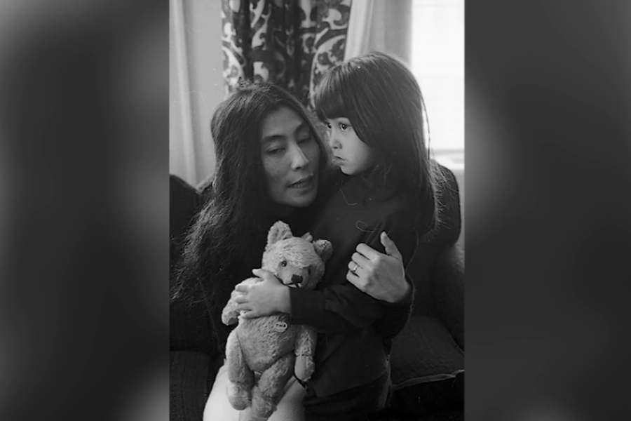 Black-and-white photo of a woman with long dark hair gently holding a young child who clutches a teddy bear. They sit on a couch in a softly lit room, with patterned curtains in the background, sharing a tender and quiet moment.