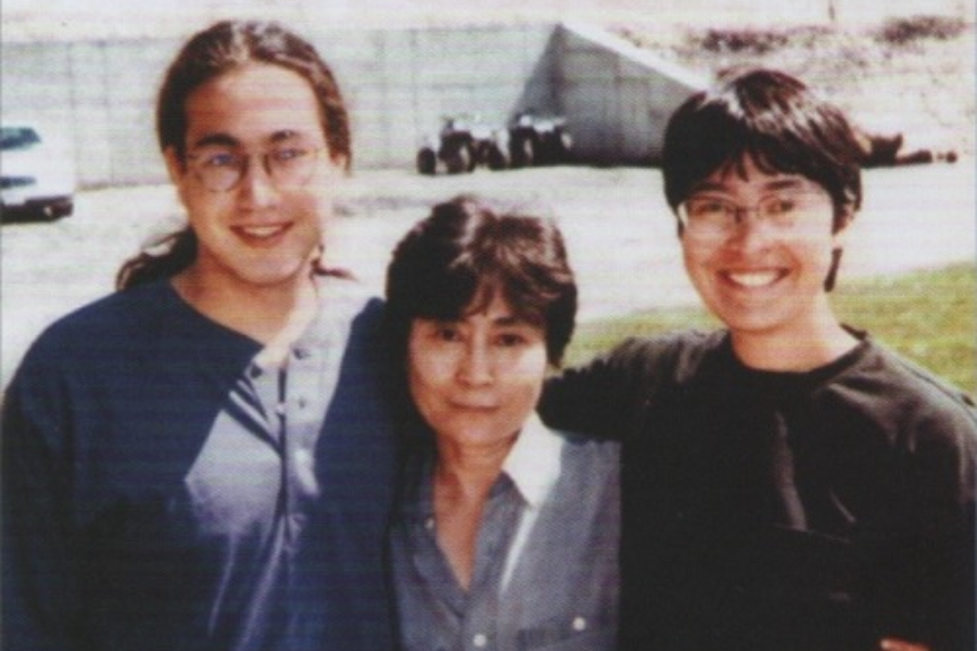 Color photo of three people standing outdoors with their arms around each other, smiling at the camera. The older woman in the middle has short dark hair and wears a blue shirt, while the two younger individuals on either side wear glasses and casual clothing, standing in front of a concrete structure.