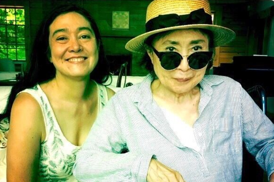 Color photo of two women indoors, one smiling brightly in a white sleeveless top, and the other wearing sunglasses, a straw hat with a black ribbon, and a light button-up shirt. They are seated together in a cozy room with wooden walls and large windows in the background.