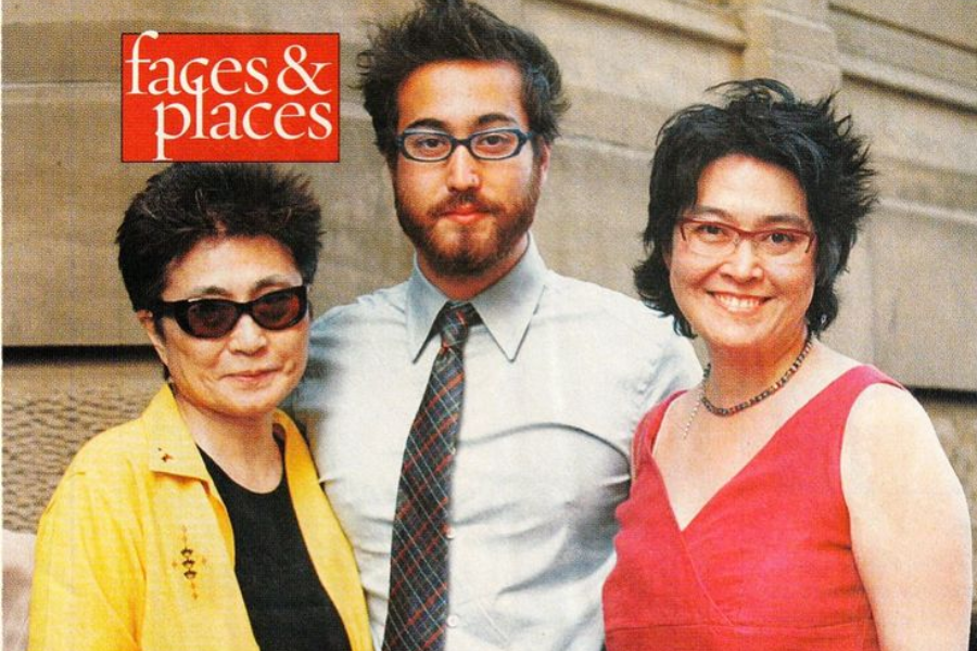 Color photo of three people posing together outdoors in front of a stone building. The woman on the left wears a yellow jacket and dark sunglasses, the man in the center has a beard, glasses, and a shirt with a tie, and the woman on the right wears a sleeveless red dress and smiles at the camera. The image features a red "faces & places" graphic in the top left corner.