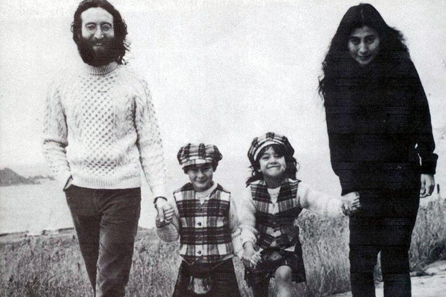 Black-and-white photo of two adults and two children walking outdoors on a grassy path. The bearded man wears a knit sweater and smiles while holding hands with one of the children in a plaid outfit, and the woman with long dark hair holds the other child’s hand as they walk together.