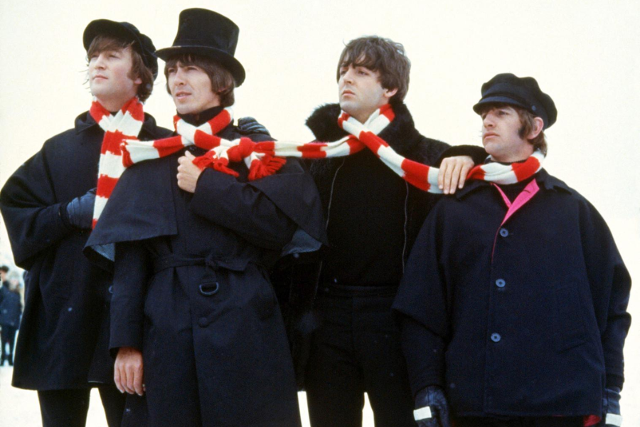 Color photo of four men standing side by side, dressed in dark winter coats with black hats, sharing a long red-and-white striped scarf wrapped around their necks. They pose against a light background, looking off in different directions.