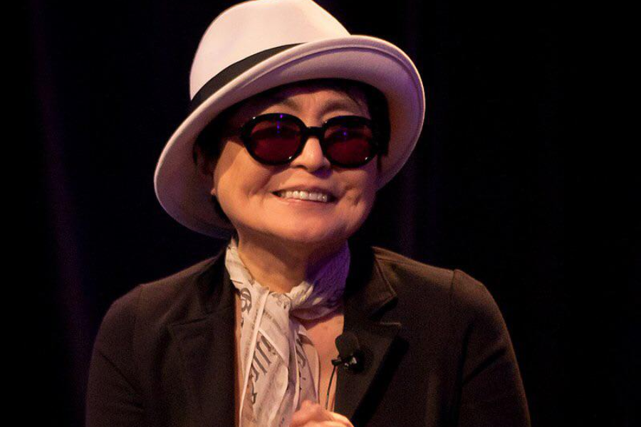 Color photo of a woman smiling while wearing a white fedora-style hat, round dark glasses with red lenses, a black jacket, and a light scarf. She is standing against a dark background, possibly on stage or at an event.