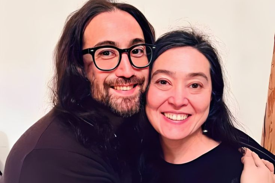 Color photo of two people with long dark hair smiling warmly as they pose together. The man on the left wears black-framed glasses and a beard, while the woman on the right has her arm around him, both standing against a plain light background.