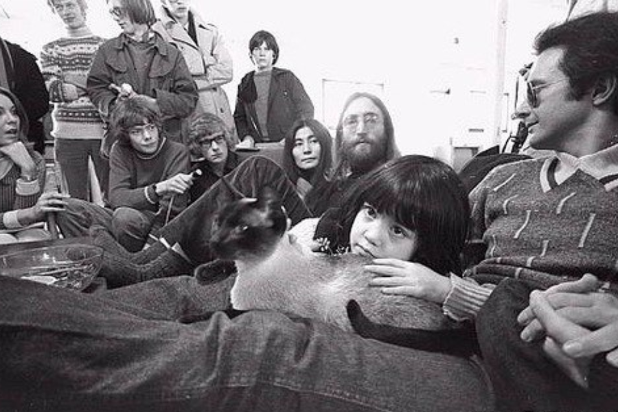 Black-and-white photo of a group of people sitting and standing closely together indoors, appearing relaxed. A child in the foreground gently holds a Siamese cat, while adults around them, including a man in glasses and a woman with long dark hair, engage in quiet conversation or look on.