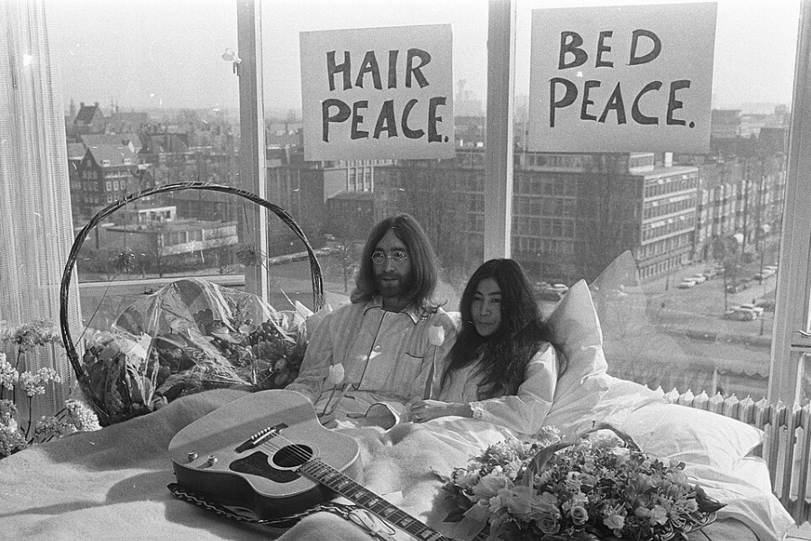Black-and-white photo of a man and woman sitting up in bed wearing pajamas during a peaceful protest, with signs reading “HAIR PEACE” and “BED PEACE” taped to the window behind them. A guitar rests on the bed alongside a bouquet of flowers, and a cityscape is visible through the window.