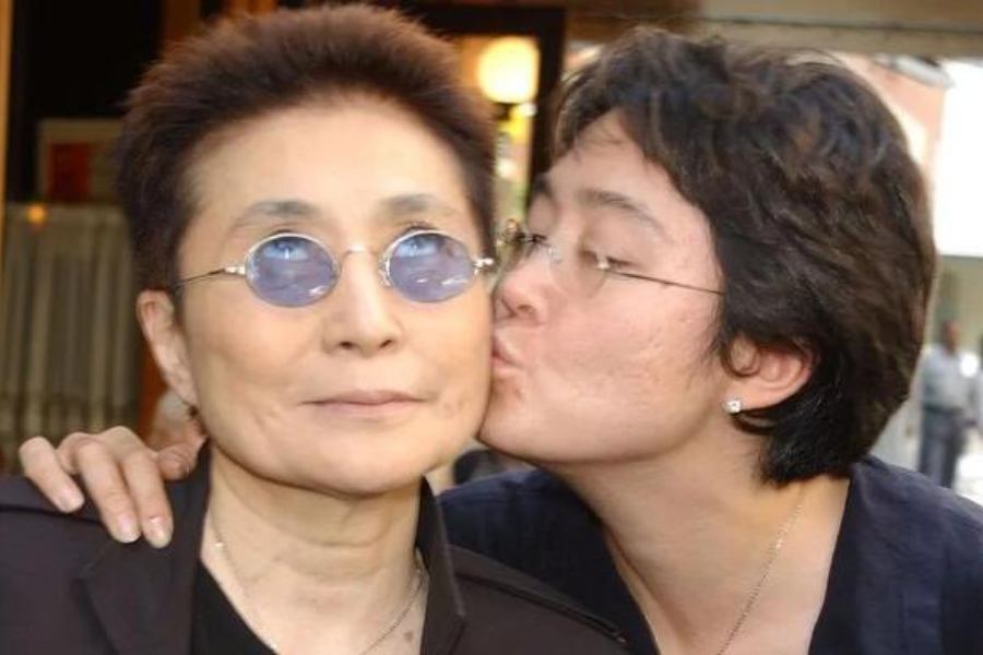 Color photo of a woman wearing round blue-tinted glasses and a black top, receiving a kiss on the cheek from another smiling woman with short dark hair and glasses. The pair is outdoors, with blurred buildings and people in the background.