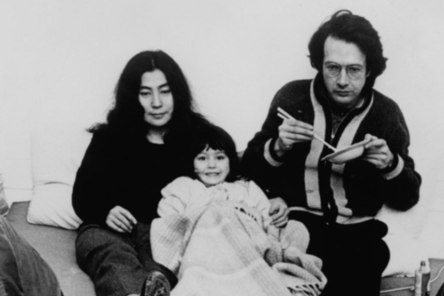 Black-and-white photo of a woman, a man, and a child sitting together on a bed or couch. The woman looks down thoughtfully, the smiling child is wrapped in a blanket, and the man wears glasses and holds chopsticks over a bowl, looking at the camera.