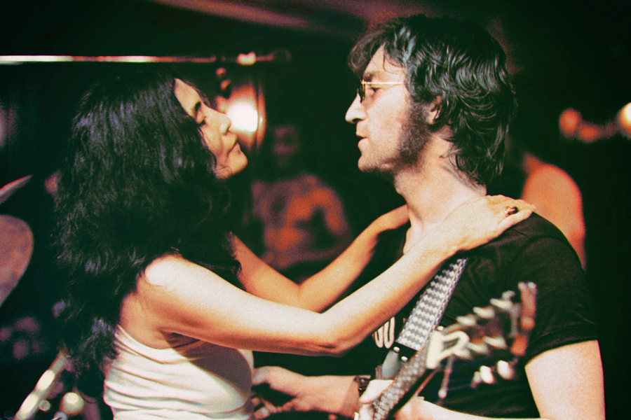 Color photo of a woman with long dark hair placing her arms around the shoulders of a man with shaggy hair and glasses, who is holding a guitar. They face each other closely in a dimly lit setting, possibly a music venue or rehearsal space.