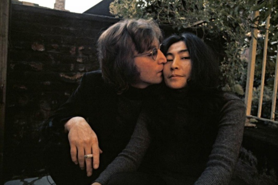 Color photograph of a man wearing glasses and holding a cigarette, gently kissing the cheek of a woman with long dark hair. They sit closely together outdoors against a brick wall with plants in the background, both dressed in dark clothing.