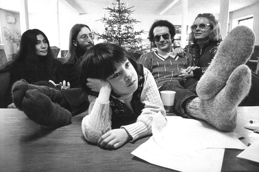 Black-and-white photo of a group of five people indoors, including a child in the foreground leaning on a table with a bored expression and wearing thick wool socks. Behind the child, two men and two women sit on a couch, relaxed, with a small Christmas tree in the background.