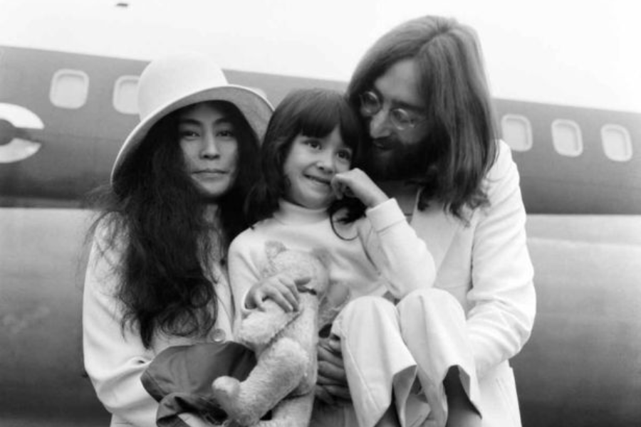 Black-and-white photo of a woman in a wide-brimmed hat standing next to a man holding a smiling young girl with a stuffed animal. They are positioned in front of an airplane, dressed in light-colored clothing.